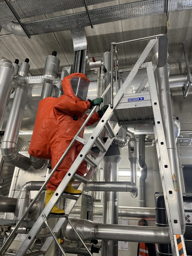 Un technicien en combinaison de sécurité orange sur une échelle en train de travailler sur une machine de froid